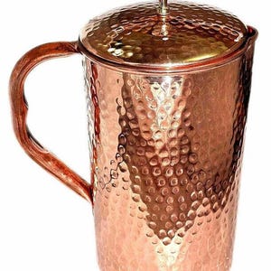 May include: A copper pitcher with a hammered texture and a matching lid. The pitcher has a curved handle and a small knob on top of the lid. The copper has a warm, reddish-brown hue.
