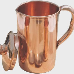 Puede incluir: Una jarra de cobre con tapa a juego. La jarra tiene un asa grande y una abertura ancha. La tapa es redonda y se coloca en el lateral. El cobre tiene una superficie brillante y reflectante.