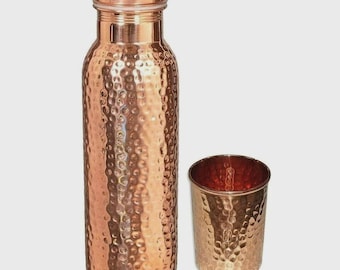 Puede incluir: Una botella de agua cilíndrica de cobre con textura martillada y tapa de rosca a juego, acompañada de un vaso de cobre martillado más pequeño. Ambos artículos presentan un acabado metálico pulido con un tono marrón rojizo cálido.