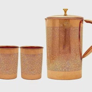 May include: A copper pitcher with a lid and handle, accompanied by two matching copper glasses. All pieces feature an etched floral pattern. The set is displayed on a white background, showcasing the warm tones of the metal.