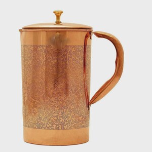 May include: A copper pitcher with a lid and handle. The pitcher has an etched floral design around the body. The handle is curved and attached to the side of the pitcher. The lid has a small knob on top.