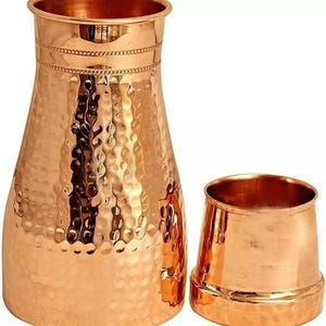Könnte beinhalten: Ein kupferfarbenes Krug- und Becher-Set. Der Krug hat einen breiten Boden, eine strukturierte Oberfläche und ein dekoratives Band in der Nähe des oberen Randes. Der Becher ist kleiner, mit einer ähnlichen gehämmerten Textur und einem ausgestellten Rand. Beide Teile haben ein glänzendes Finish.
