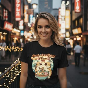 May include: A black short-sleeved t-shirt featuring a graphic of a ginger cat wearing a white headband and eating ramen from a blue bowl with chopsticks. Above the cat, Japanese text is printed in a curved orange banner against a teal circle.