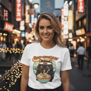 May include: A white short-sleeved t-shirt featuring a graphic of a black cat wearing a headband and eating ramen from a bowl with chopsticks. Above the cat, the Japanese text reads "我のラーメンに触るな!" set against a circular teal background with wave patterns.