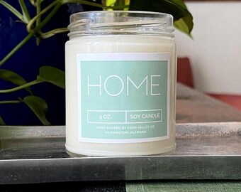 May include: A clear glass jar candle with a white wax interior. The label reads "HOME" in white letters on a light teal background. It is a 9 oz soy candle, hand-poured in Hamilton, Alabama.