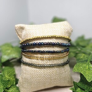 May include: Several beaded bracelets in shades of gold, dark blue, and clear are presented on a small burlap cushion. The bracelets are composed of tiny, spherical beads. Green leaves frame the cushion, providing a natural aesthetic.
