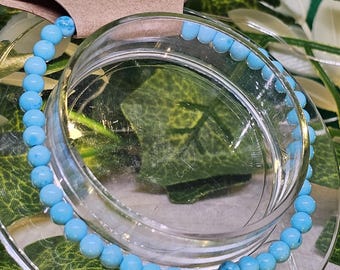 May include: A delicate bracelet featuring small, round, light blue beads. The beads are strung together, forming a complete circle. The bracelet is displayed on a clear glass surface, with a natural green background.