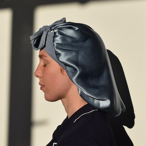 May include: A dark teal satin bonnet with a matching bow, designed to protect hair. The bonnet is worn over a light grey cap. The person is wearing a navy blue top with white trim. The background is a light beige colour.