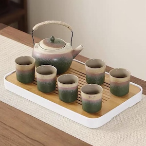 May include: A ceramic tea set comprising a teapot and six cups. The teapot features a woven handle and a lid with a small knob. The cups and teapot have a green, pink, and beige colour scheme. The set is presented on a bamboo tray.