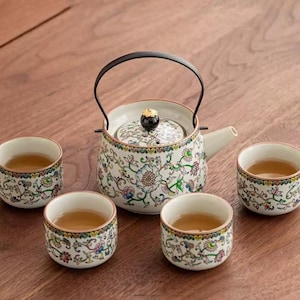Puede incluir: Juego de té de cerámica con una tetera y cuatro tazas a juego. La tetera tiene un asa negra y está decorada con motivos florales en azul, verde y rosa. Las tazas y la tetera están llenas de té.