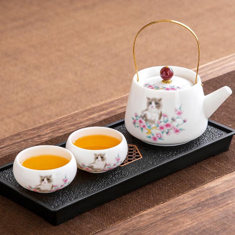 Op de afbeelding: Een witte keramische theeset met een kattenontwerp en roze bloemaccenten. De set bevat een theepot met een gouden handvat en twee theekopjes gevuld met thee, gepresenteerd op een zwarte dienblad. De theepotdeksel heeft een rode knop.