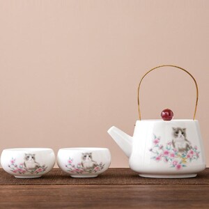 Op de afbeelding: Een witte keramische theeset met een theepot en twee kopjes. Elk stuk is versierd met een aquarel illustratie van een kat omringd door roze bloemen. De theepot heeft een gouden handvat en een rode knop.