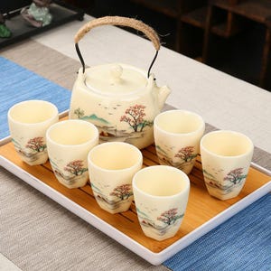 Op de afbeelding: Een crèmekleurige keramische theeset met een theepot en zes bijpassende kopjes, allemaal versierd met landschapsschilderijen met bomen en bergen. De theepot heeft een geweven handvat. De set is gerangschikt op een bamboe dienblad.