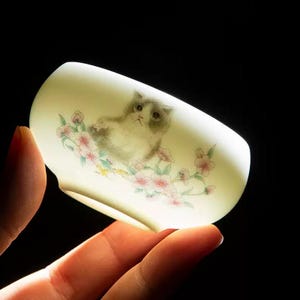 Op de afbeelding: Een kleine, cr&egrave;mekleurige keramische theekop met een handgeschilderde illustratie van een grijs-witte kat te midden van roze bloesems. De kop heeft een afgeronde vorm en wordt tegen een donkere achtergrond gehouden.