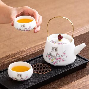 Op de afbeelding: Een witte keramische theeset met een theepot, een gouden handvat en een rode knop op het deksel, en twee theekopjes. Elk stuk is versierd met een kattenillustratie en roze bloemaccenten. De kopjes zijn gevuld met thee.