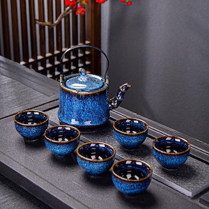 Puede incluir: Un juego de té de cerámica azul con una tetera y seis tazas a juego. La tetera tiene un asa negra y un pico decorativo. Las tazas y la tetera tienen un esmalte moteado con ribete marrón. El juego está dispuesto sobre una superficie gris oscura.