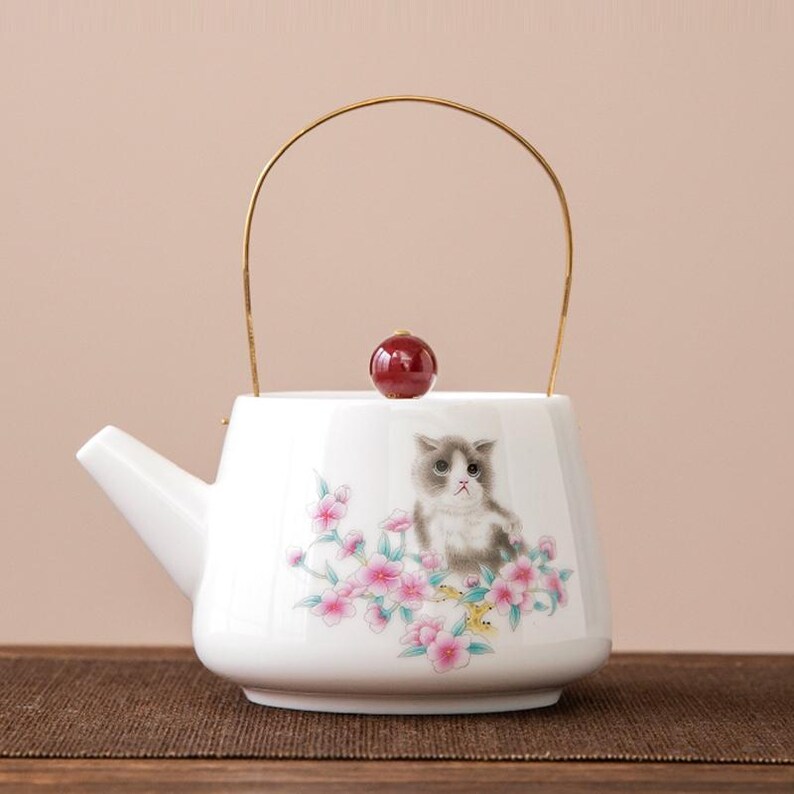 Op de afbeelding: Witte keramische theepot met een gouden handvat en een rode knop op het deksel. De theepot heeft een geschilderd ontwerp van een grijze en witte kat omringd door roze bloemen. De tuit bevindt zich aan de linkerkant.