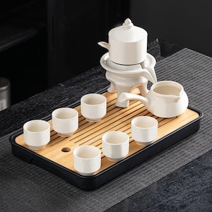 Può includere: Un set da tè in ceramica bianca con un vassoio di bambù. Il set include una teiera, una brocca, sei tazzine e un infusore per il tè. Il vassoio è rettangolare con un bordo nero. Il set da tè è progettato per servire il tè.