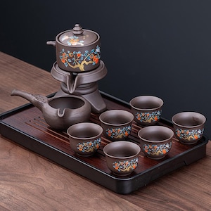 May include: A brown ceramic tea set with a teapot, server, and six cups, all decorated with a floral design. The set sits on a dark wooden tray. The teapot has a lid and a spout. The cups are small and round.