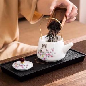 Op de afbeelding: Witte keramische theepot met een kattenillustratie en bloemaccenten, met een gouden handvat. De theepot staat op een zwarte tray met een bijpassende deksel. Theebladeren worden erin gegoten.