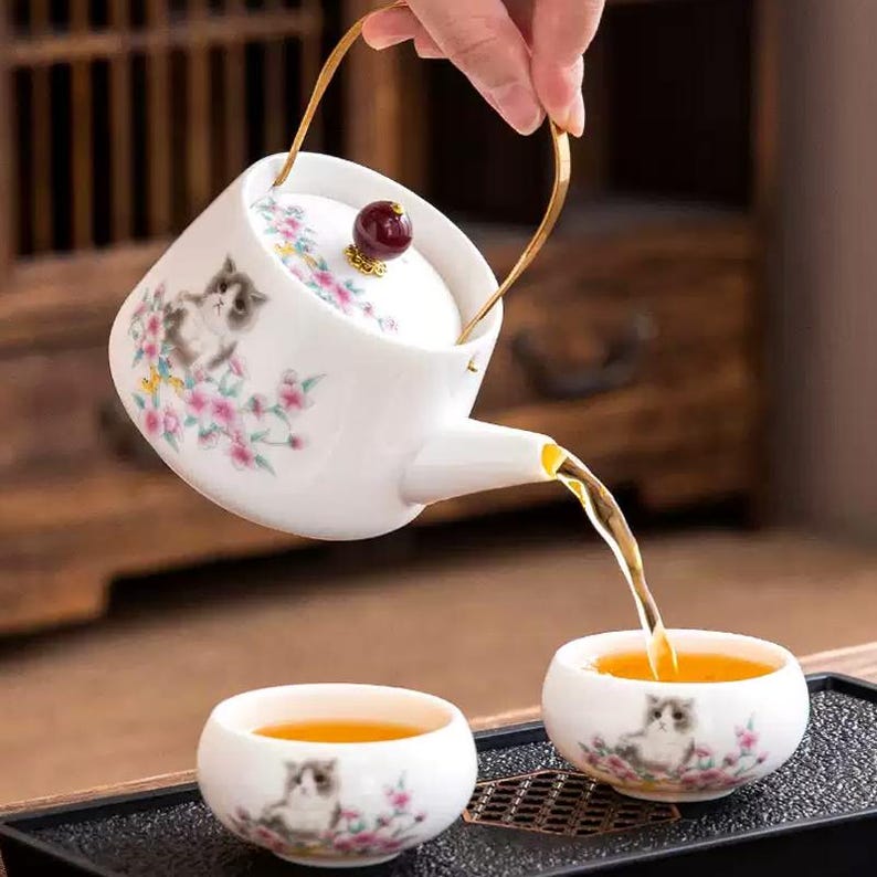 Op de afbeelding: Witte keramische theepot en theekopjes met katten- en bloemmotieven. De theepot heeft een gouden handvat en schenkt thee in een theekopje. De thee heeft een gouden kleur. De theekopjes en de theepot staan op een zwart dienblad.