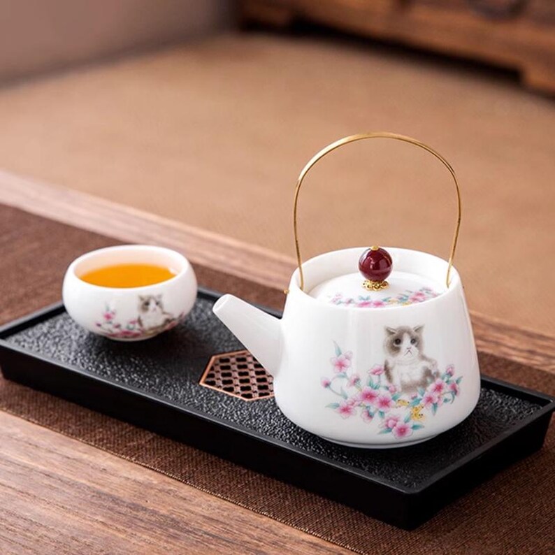 Op de afbeelding: Witte keramische theepot en kopjes set met een kattenillustratie en bloemenaccenten. De theepot heeft een gouden handvat en een rode knop op het deksel. De set staat op een zwarte tray.