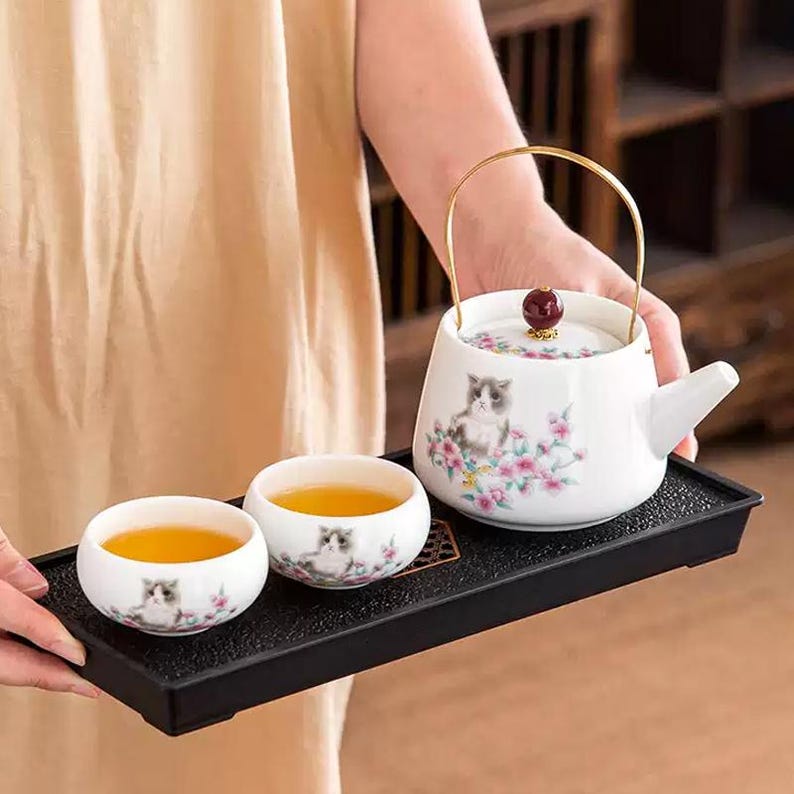 Op de afbeelding: Een witte keramische theeset met een katten- en bloemenontwerp, inclusief een theepot met een gouden handvat en een rode knop, en twee kopjes. De set wordt gepresenteerd op een zwarte dienblad.
