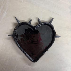Può includere: Un vassoio nero a forma di cuore con finitura lucida e bordo rialzato. Il vassoio è adornato da diverse punte grigio scuro lungo il perimetro. Il vassoio è su una superficie di legno chiaro.