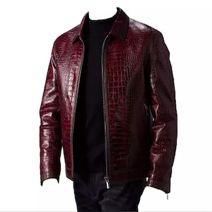 Puede incluir: Chaqueta de cuero burdeos con textura de piel de cocodrilo. La chaqueta tiene cierre de cremallera, cuello y mangas largas. La chaqueta se lleva sobre un cuello alto negro.
