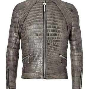 Peut inclure: Veste en cuir grise avec un motif texturé en forme de crocodile. La veste est dotée d'un col montant, d'une fermeture éclair sur toute la longueur et de poches zippées sur la poitrine et les manches. Les manches ont des fermetures éclair aux poignets.