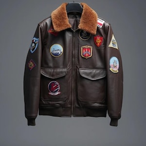 May include: A brown leather bomber jacket with a tan fur collar. The jacket has several embroidered patches on the chest and sleeves. It features two front pockets and ribbed cuffs. The jacket is suspended on a black hanger against a grey backdrop.