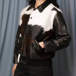 May include: A black and white cowhide jacket with black leather sleeves, collar, and trim. The jacket has a button-up front, two pockets, and a ribbed hem. The jacket is a stylish outerwear piece.