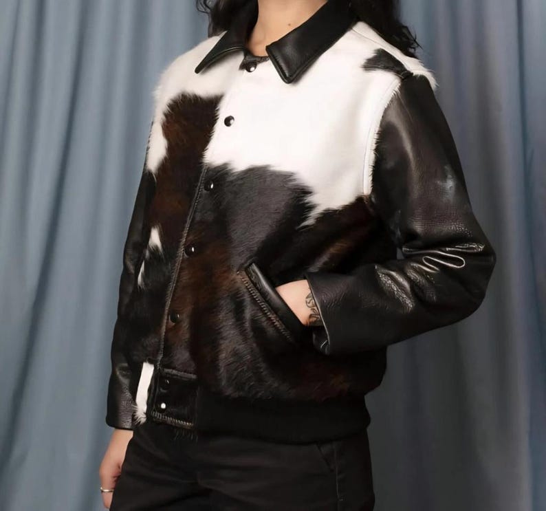 May include: A black and white cowhide jacket with black leather sleeves, collar, and trim. The jacket has a button-up front, two pockets, and a ribbed hem. The jacket is a stylish outerwear piece.