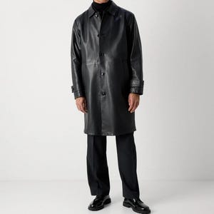 May include: A black leather coat with a button-up front, long sleeves, and a collar. The coat is knee-length and worn with black pants and shoes. The coat has a classic, tailored design.