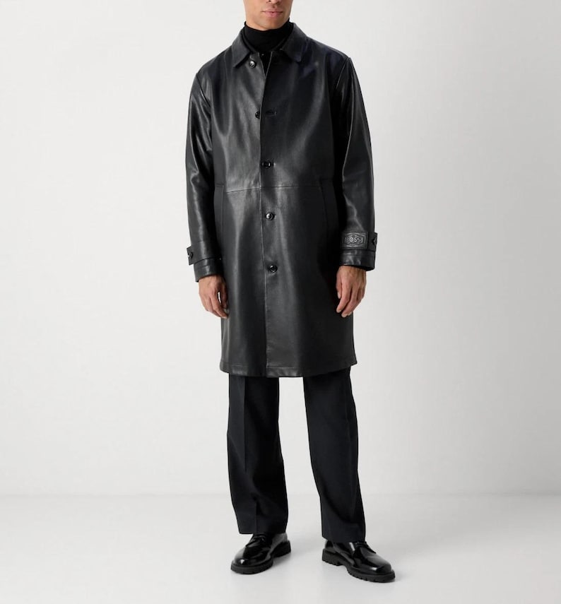 May include: A black leather coat with a button-up front, long sleeves, and a collar. The coat is knee-length and worn with black pants and shoes. The coat has a classic, tailored design.