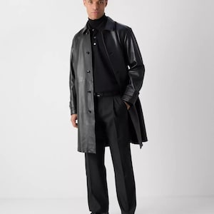 May include: A black leather trench coat with a button-up front, worn over a black polo shirt and black trousers. The outfit is completed with black dress shoes. The model is standing in front of a white background.