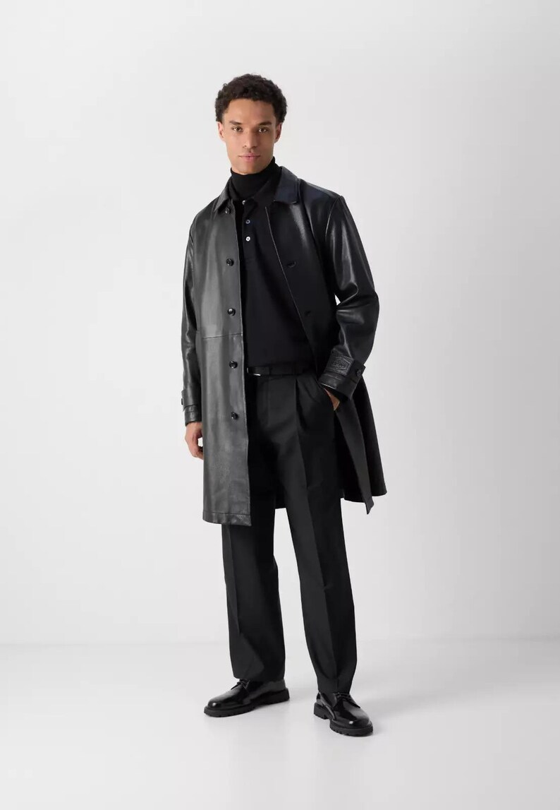 May include: A black leather trench coat with a button-up front, worn over a black polo shirt and black trousers. The outfit is completed with black dress shoes. The model is standing in front of a white background.
