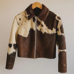 May include: A brown and cream-coloured jacket with a cowhide pattern. The jacket has a collar, a full-length zip, and long sleeves. The jacket is hanging on a wooden hanger.