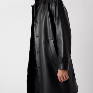 May include: A black leather trench coat with a button-up front, a collar, and a tailored fit. The coat features a sleek, minimalist design, with a length that extends to the mid-calf. The coat has a classic, timeless style.