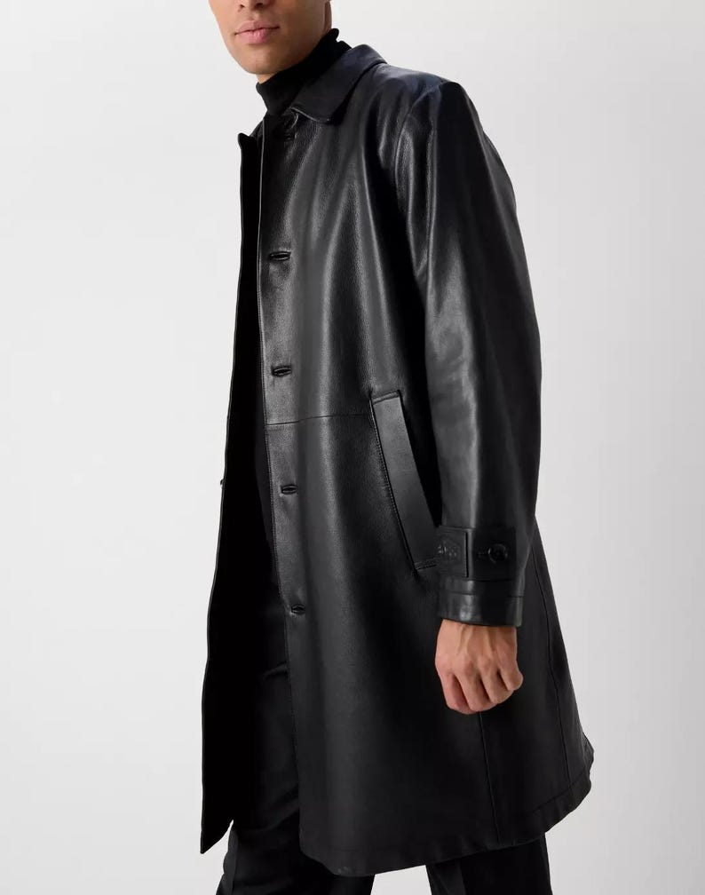 May include: A black leather trench coat with a button-up front, a collar, and a tailored fit. The coat features a sleek, minimalist design, with a length that extends to the mid-calf. The coat has a classic, timeless style.