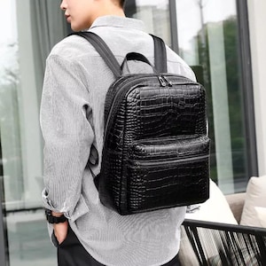 May include: Black backpack with a crocodile skin texture. The backpack has a top handle, adjustable shoulder straps, and a front zippered pocket. The bag is worn on the back.