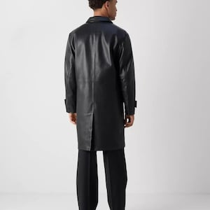 May include: A black leather coat with a classic design. The coat is long, reaching below the knees, and features a notched collar and button closures. The model is wearing black pants and shoes.