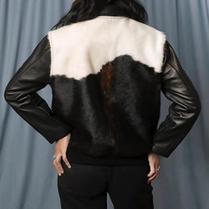 May include: A black leather jacket with a white and black cowhide back panel. The jacket has long sleeves and a round collar. The model is wearing black pants.