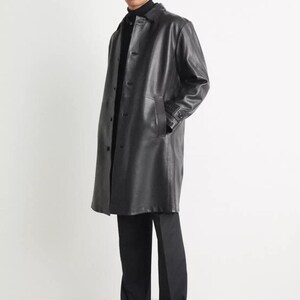 May include: A black leather coat with a button-up front, worn over a black turtleneck and black pants. The coat has a classic design and reaches to mid-calf. The model is wearing black dress shoes.