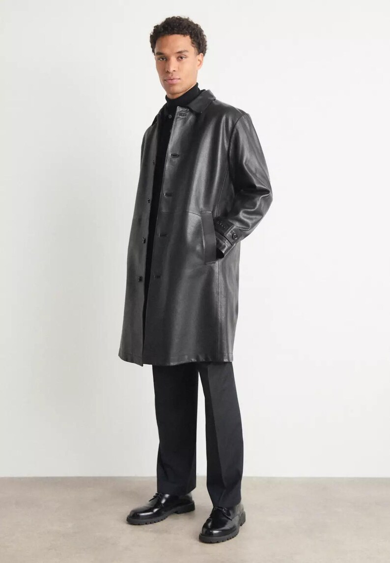 May include: A black leather coat with a button-up front, worn over a black turtleneck and black pants. The coat has a classic design and reaches to mid-calf. The model is wearing black dress shoes.