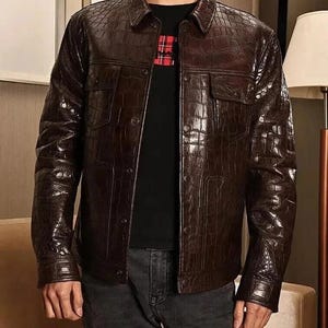 Może przedstawiać: Brązowa skórzana kurtka o fakturze skóry krokodyla. Kurtka ma dwie kieszenie na piersi, zapięcie na guziki i kołnierz. Noszona na czarnym t-shircie i ciemnoszarych dżinsach.