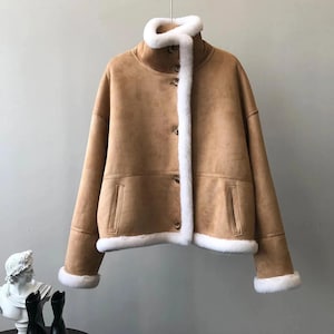 Może przedstawiać: Jasnobrązowa zamszowa kurtka z wysokim kołnierzem i białym wykończeniem z imitacji futra. Kurtka ma zapięcie na guziki, dwie kieszenie i białe wykończenie na mankietach i na dole. Stylowa odzież wierzchnia.