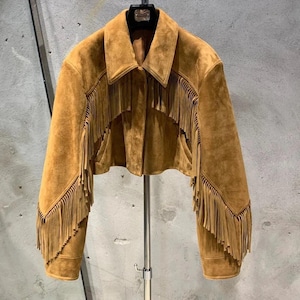 Puede incluir: Chaqueta de ante marrón claro con mangas largas y cuello. La chaqueta presenta flecos decorativos en la parte delantera, las mangas y la espalda. Cuelga de una percha metálica.