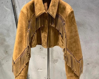 May include: A tan suede jacket with a collar and long sleeves. The jacket has decorative fringe detailing on the front, sleeves, and back. It is displayed on a metal clothes rack.