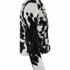 May include: A side view of a moto-style jacket featuring a black and white cow-print pattern. The jacket includes a notched collar, long sleeves with zippered cuffs, a silver-toned front zipper, and a buckled belt detail at the waist hem.